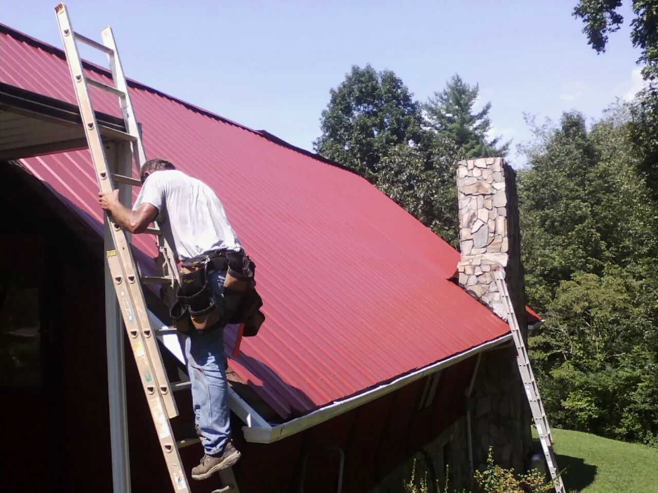 Roofer on a ladder beside a red metal roof and stone chimney — Soffit & Fascia Repair by Roofing Brothers in Lockeford